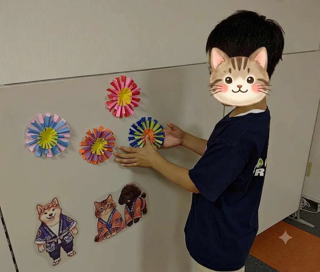帯広東教室 展示活動 — 完成した色紙の花飾りを教室の壁に自分で選んで貼る子ども、下に柴犬・猫・小犬の祭りマスコットも並ぶ（帯広市 学習支援特化型 児童発達支援・放課後等デイサービス）