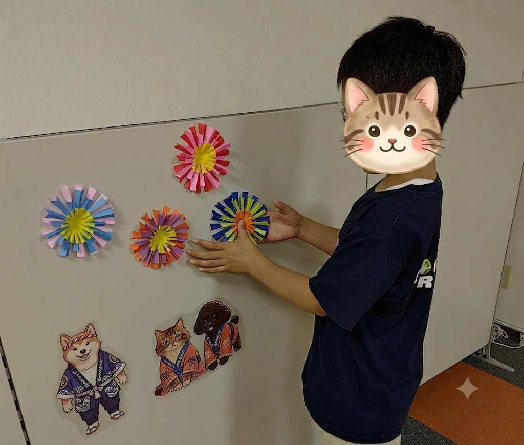 帯広東教室 展示活動 — 完成した色紙の花飾りを壁に貼る子どもの様子、周りに祭りマスコットも並ぶ